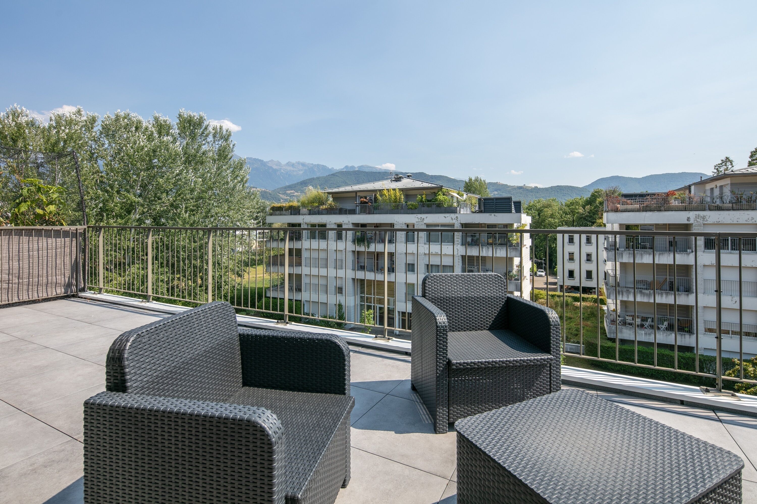 A rooftop terrace with wicker furniture and a view of buildings and mountains.