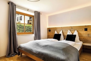 A bedroom with a large bed, grey curtains, a wooden headboard, and a window with a view of greenery. - KitzAlps Standard Apartment (Kirchberg in Tirol)