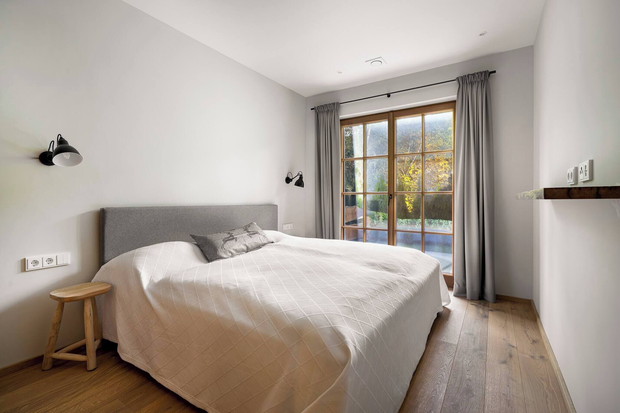 Une chambre à coucher avec un grand lit, un tabouret en bois, une fenêtre avec des rideaux et des lampes fixées au mur.