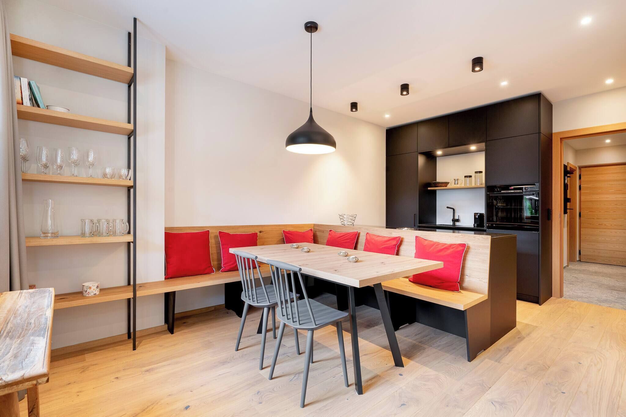 Una cocina moderna con horno empotrado, un comedor con mesa de madera y sillas grises, y una estantería con cristalería.