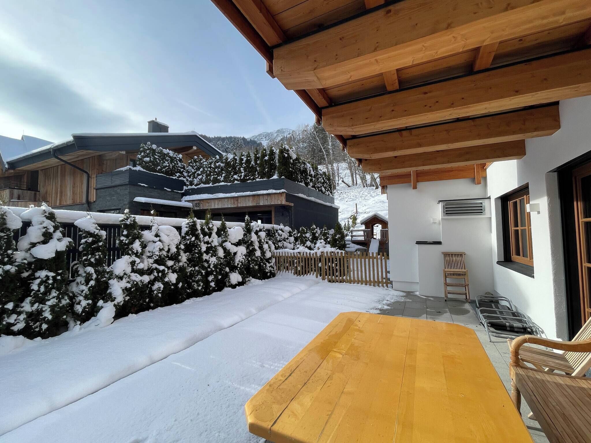 Une terrasse en bois avec une table et des chaises, entourée d’arbres couverts de neige et d’un bâtiment doté d’un toit en pente.