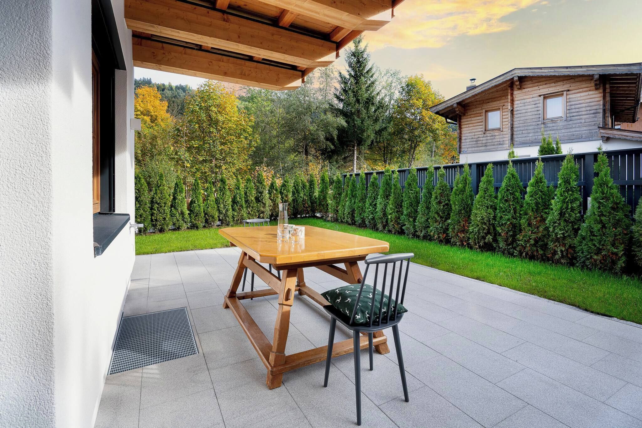 Una terrazza esterna con tavolo e sedie in legno, un barbecue in metallo e vista su una casa in legno e un siepe.