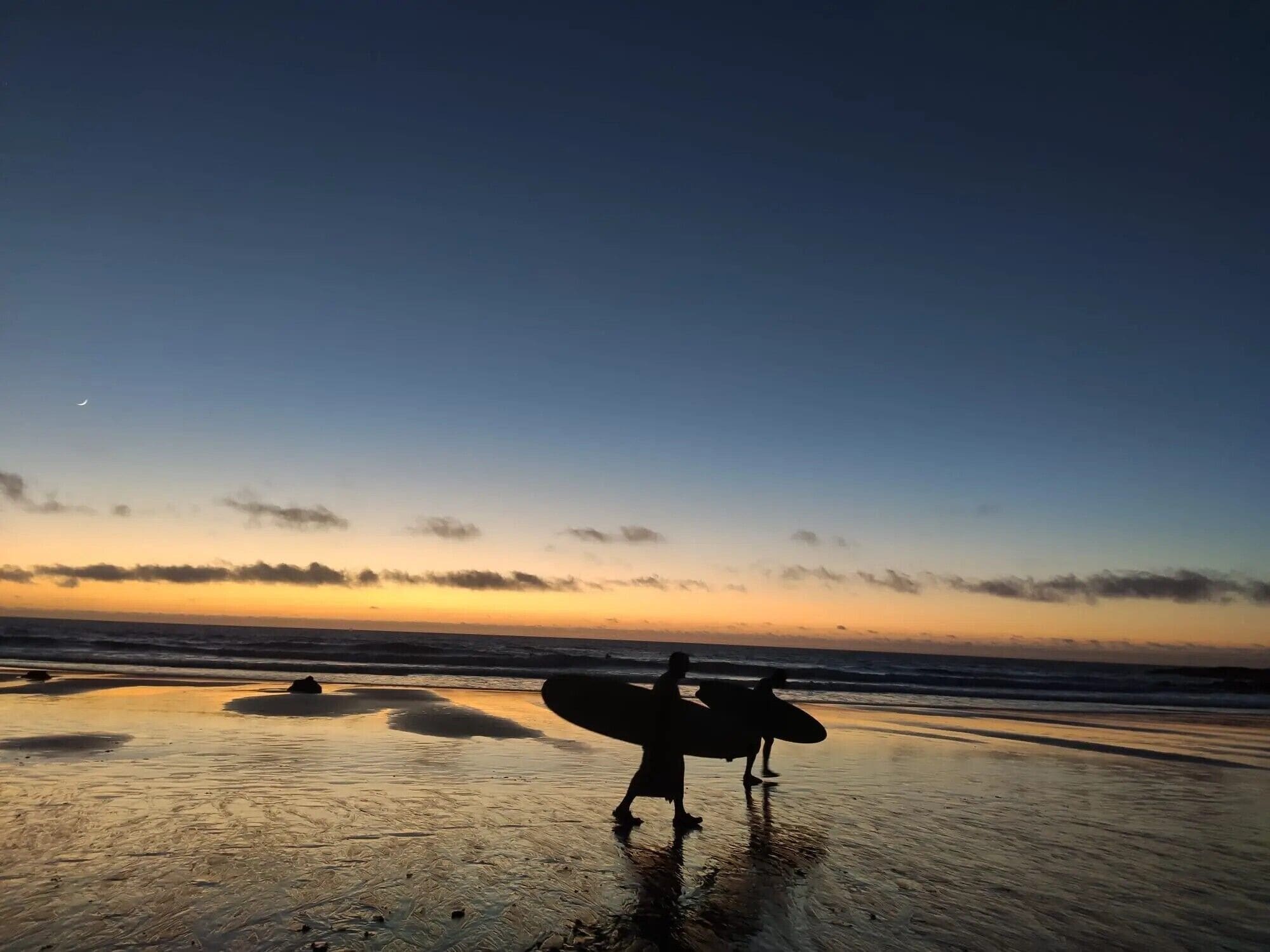 Escena de playa con una persona sosteniendo una tabla de surf, olas y una puesta de sol.