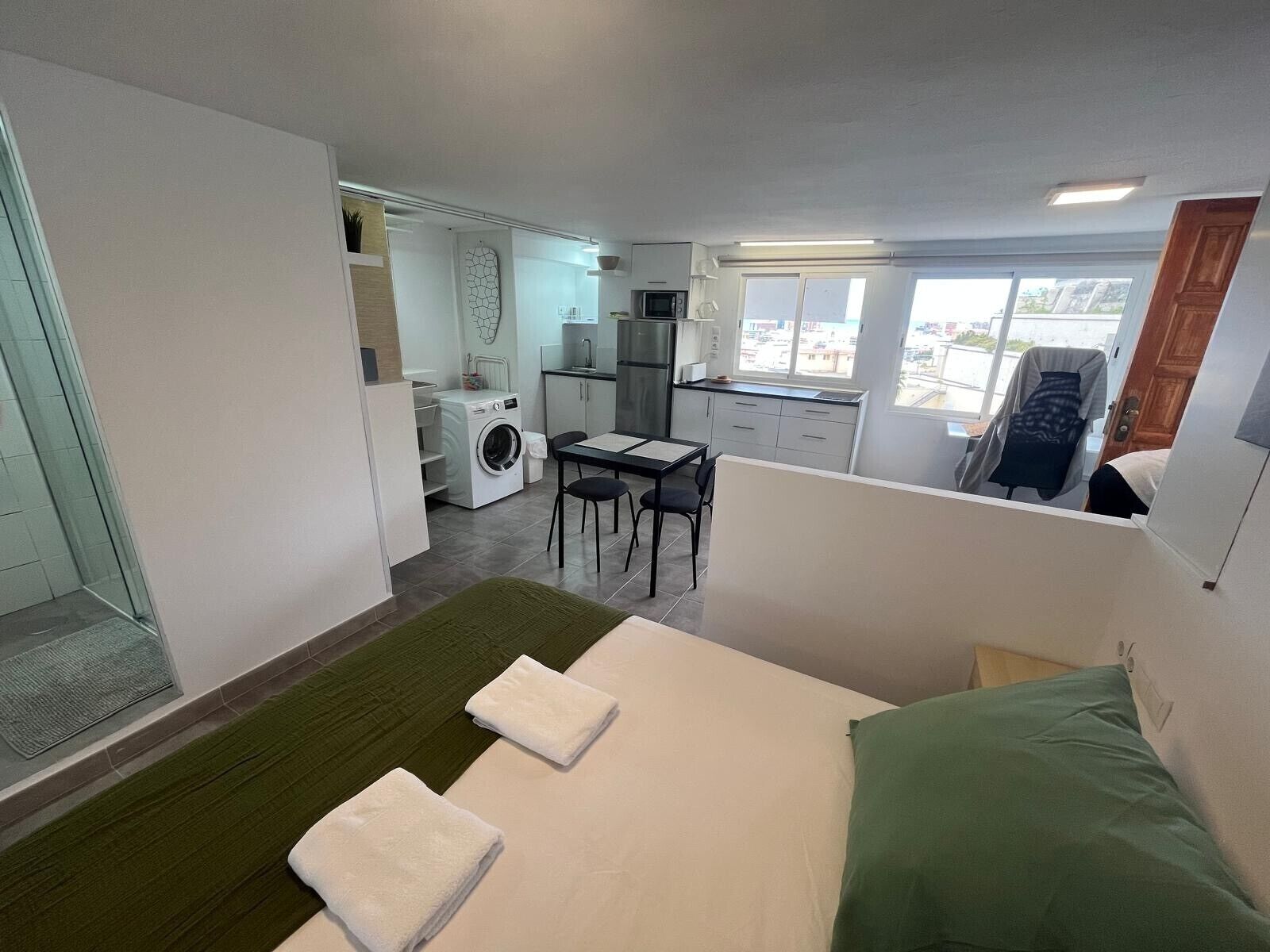 A hotel room with a bed, a small table, a chair, a washing machine, and a kitchenette.