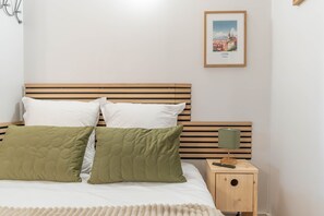 Un lit bien fait, avec une tête de lit en bois, deux oreillers, un oreiller vert matelassé et un oreiller blanc. Un tableau encadré représentant Lyon, en France, est accroché au mur.