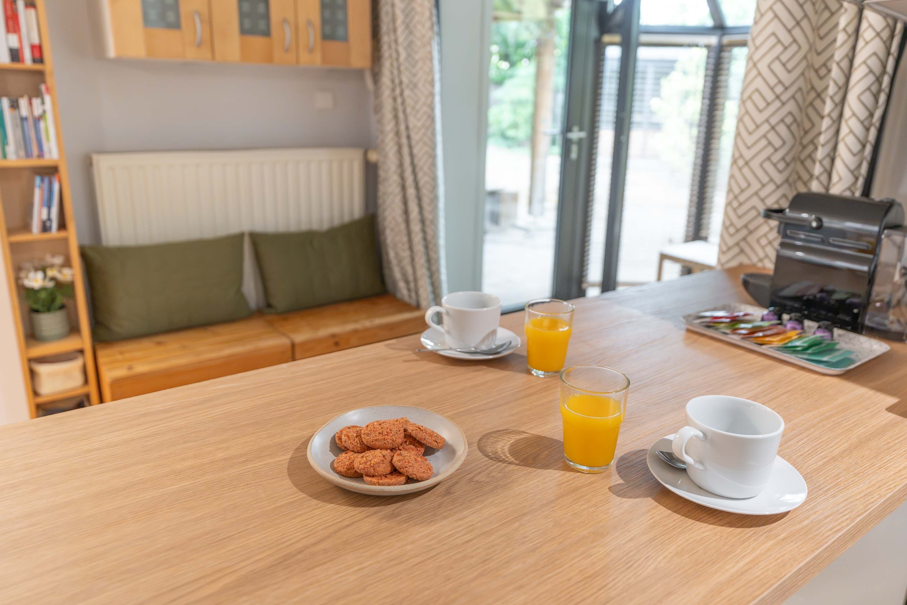 Una preparación para el desayuno con un plato de galletas, dos vasos de jugo de naranja y dos tazas blancas sobre una mesa de madera.