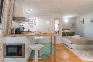 A compact hotel room with a kitchenette, a bed with a green pillow, and a small bathroom. - Le Cottage - Studio avec véranda et jardin (Lyon)