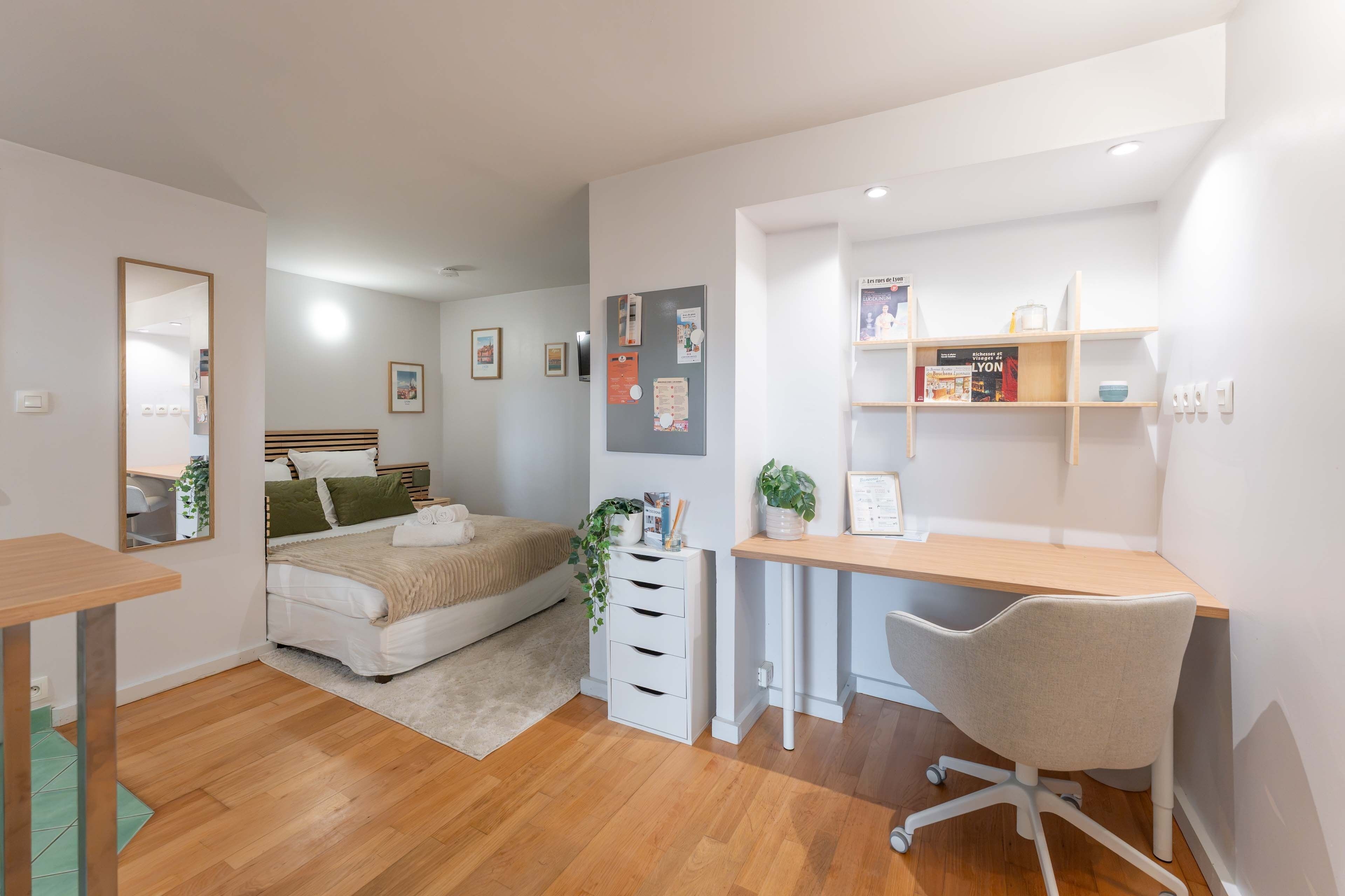 Quarto moderno com uma cama, mesa de cabeceira e escrivaninha com cadeira. Há livros e uma planta na escrivaninha.