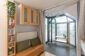 A cozy reading nook with a wooden bench, a bookshelf, and a view through a glass door to an outdoor area. - Le Cottage - Studio avec véranda et jardin (Lyon)
