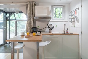 A compact kitchen with a wooden table, white chairs, a window, and a built-in oven. - Le Cottage - Studio avec véranda et jardin (Lyon)