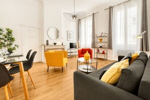 Ein modernes Wohnzimmer mit einer Couch, einem Esstisch mit Stühlen, einem Fernseher und einem großen Fenster mit Vorhängen.