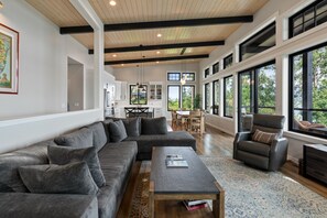 A spacious living room with a large gray sectional sofa, a wooden coffee table, and a view of the outdoors through large windows. - Luxury Lakefront Retreat | Private Beach Near Town (Sagle)