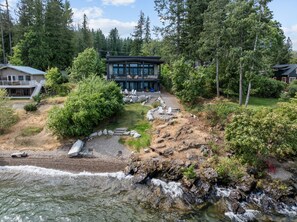 A modern house with a large deck, surrounded by dense forest and a rocky shoreline. - Luxury Lakefront Retreat | Private Beach Near Town (Sagle)