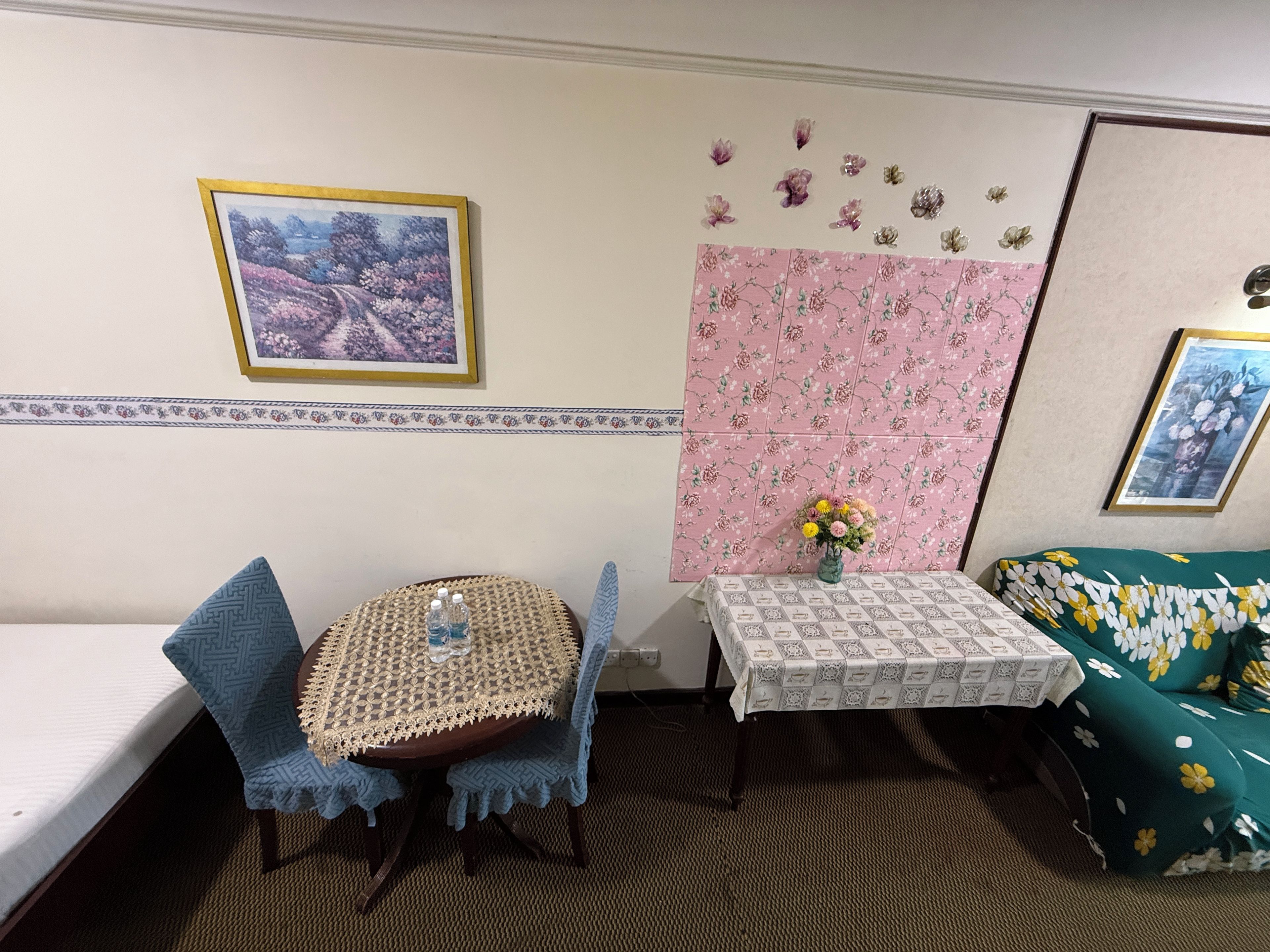 Una habitación con una cama, una pequeña mesa redonda con mantel de encaje, dos sillas, un sofá con funda estampada de flores, una pared con diseño floral, y un cuadro enmarcado en la pared.