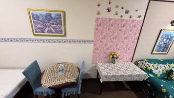Una habitación con una cama, una pequeña mesa redonda con mantel de encaje, dos sillas, un sofá con funda estampada de flores, una pared con diseño floral, y un cuadro enmarcado en la pared.