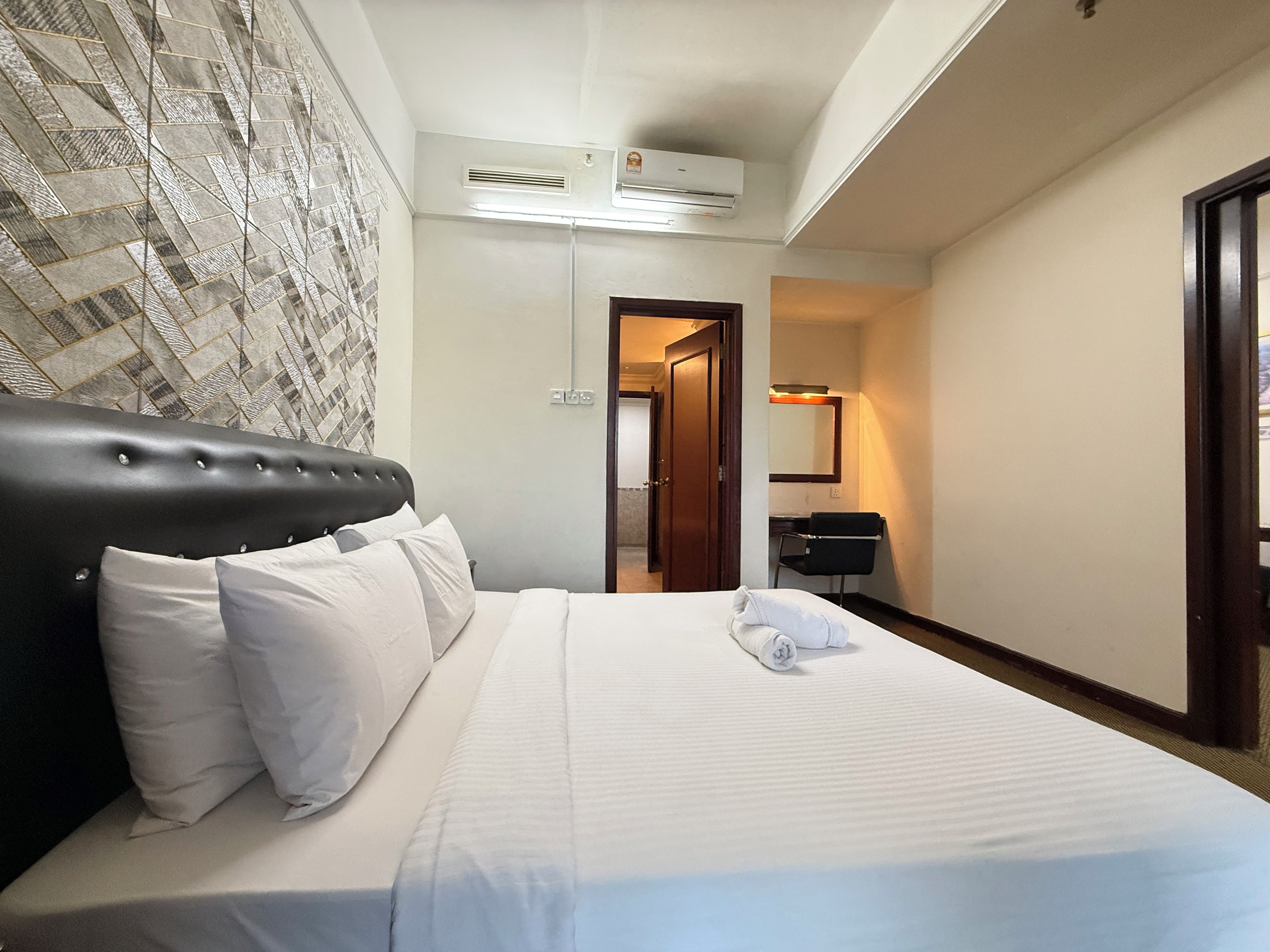 A hotel room with a bed, a wall-mounted air conditioner, a door leading to a bathroom, and a desk with a chair.
