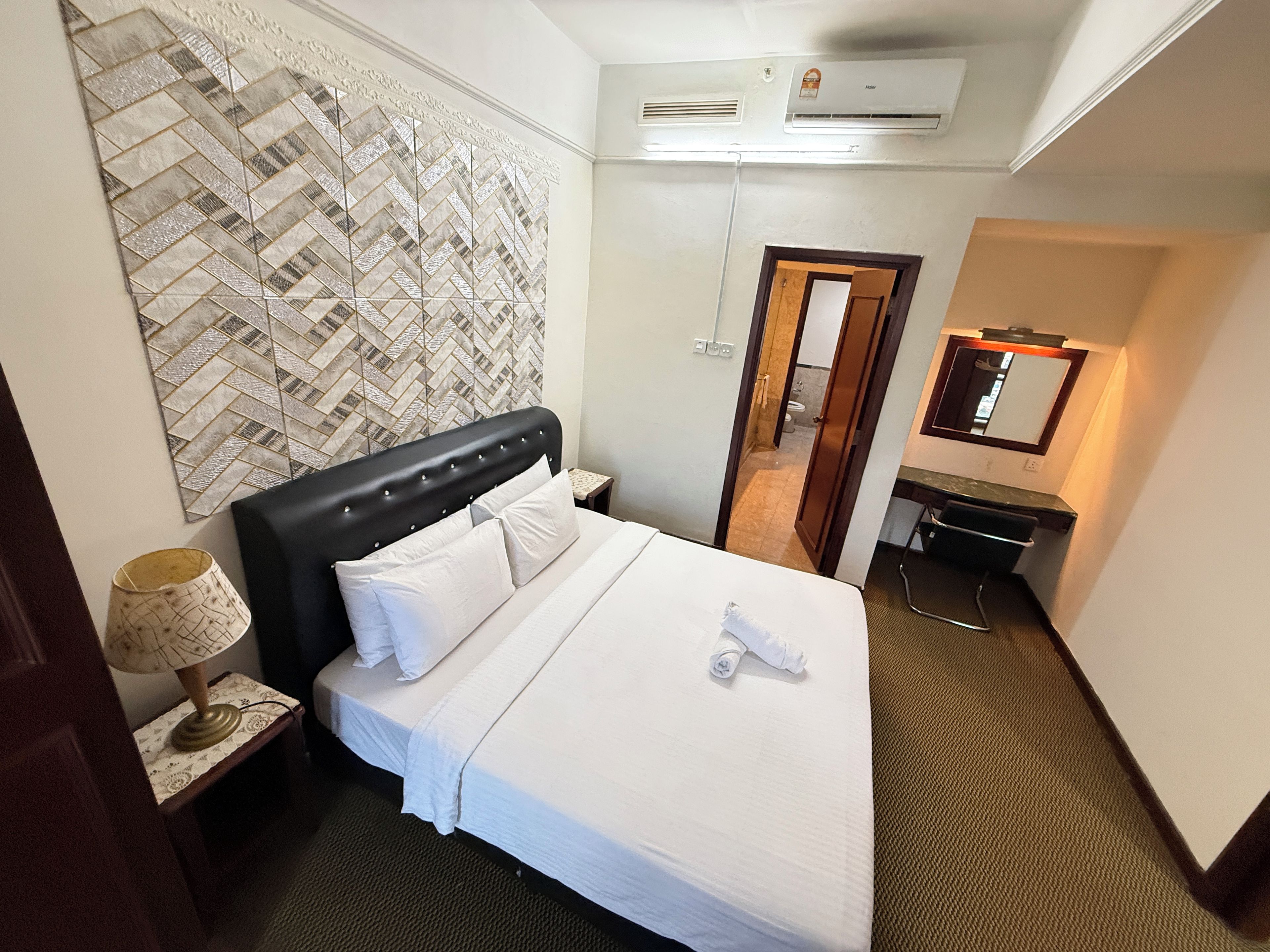 Habitación de hotel con cama, mesita de noche, lámpara, espejo y baño visible a través de una puerta abierta.