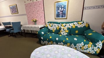 A room with a floral sofa, a dining table with chairs, a small table with a vase of flowers, and a framed picture on the wall.