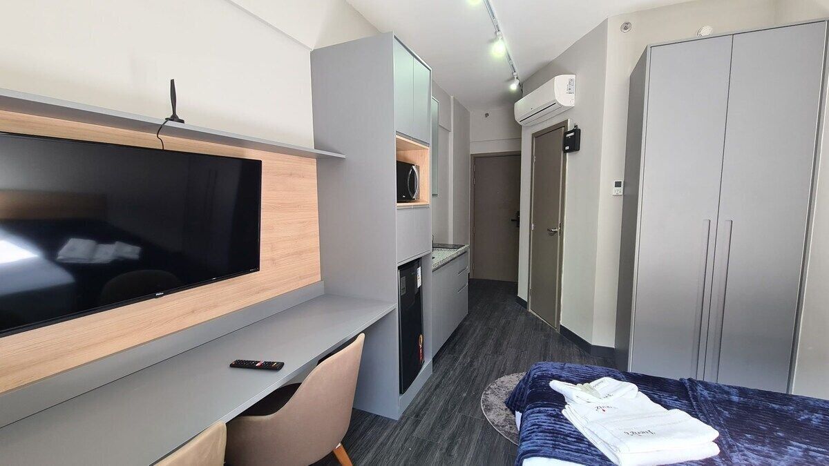Habitación de hotel moderna con televisión de pantalla plana, escritorio, silla, cama con manta azul y armario.