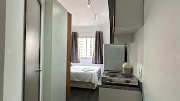 Habitación de hotel con cama, kitchenette pequeña, espejo y ventana con cortinas.