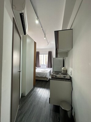 Habitación de hotel con cama, kitchenette pequeña, espejo y ventana con cortinas.