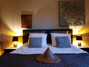 Una cama con ropa blanca, dos almohadas, un tapete tejido y dos lámparas de noche.