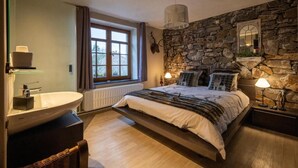 Een slaapkamer met een stenen muur, een bed met meerdere kussens, een nachtkastje met een lamp en een raam met gordijnen.