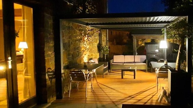 Een terras buiten met een eettafel en stoelen, een stenen muur, en een voertuig op de achtergrond.