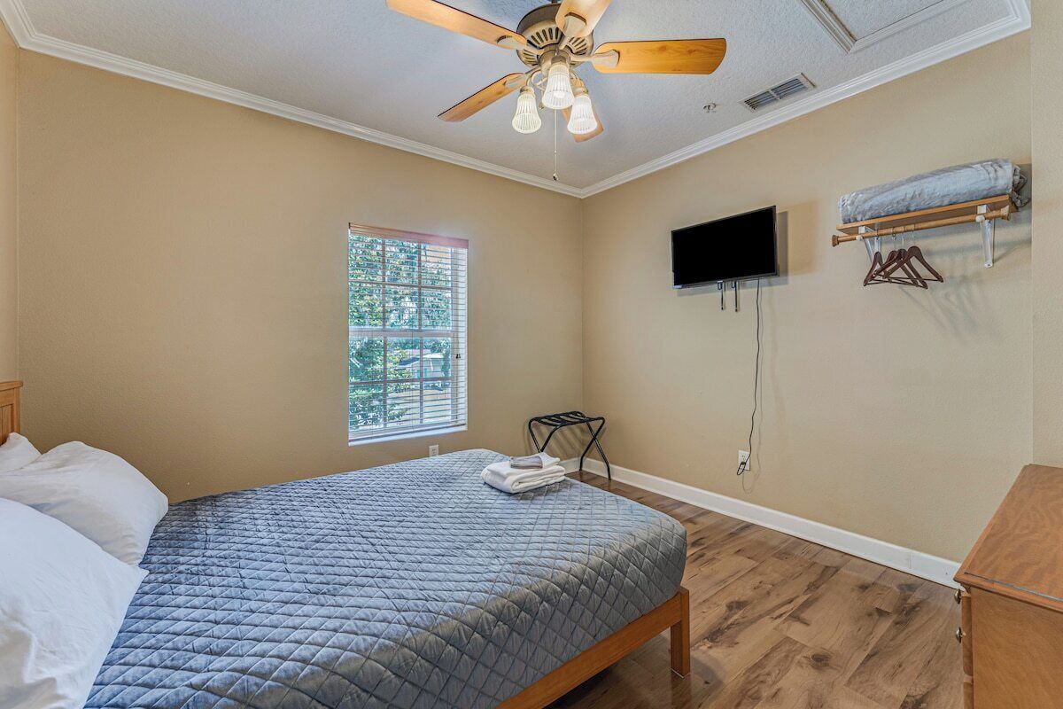 A bedroom with a bed, a ceiling fan, a TV mounted on the wall, and a window with blinds.