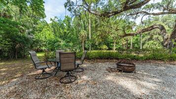 An outdoor seating area with a table and chairs, surrounded by trees and a gravel ground.