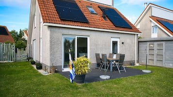 Une maison avec un toit en pente, des panneaux solaires, une terrasse avec une table et des chaises, et un drapeau.