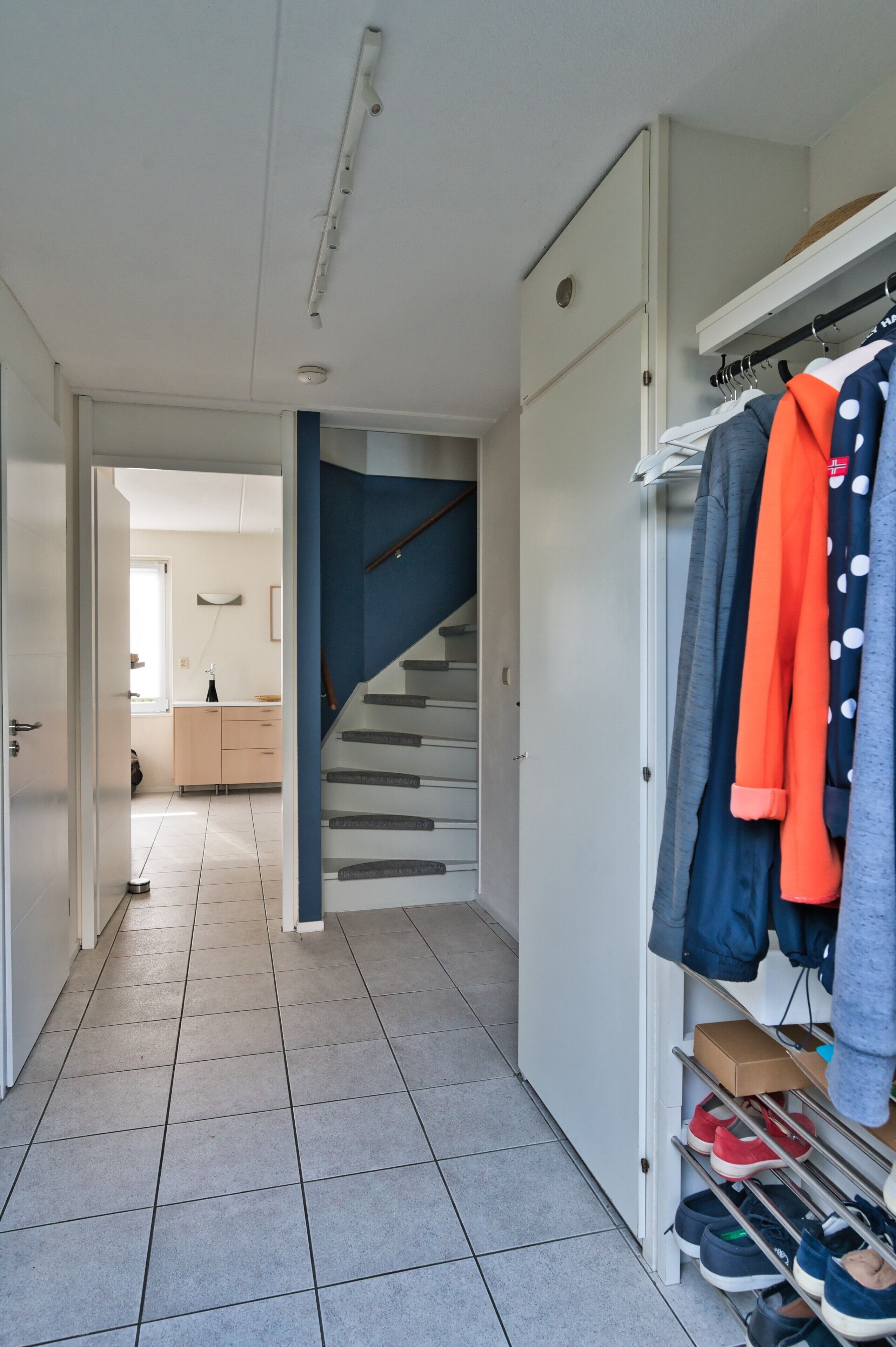 A hallway with a tiled floor, a staircase, and a clothing rack with various garments.