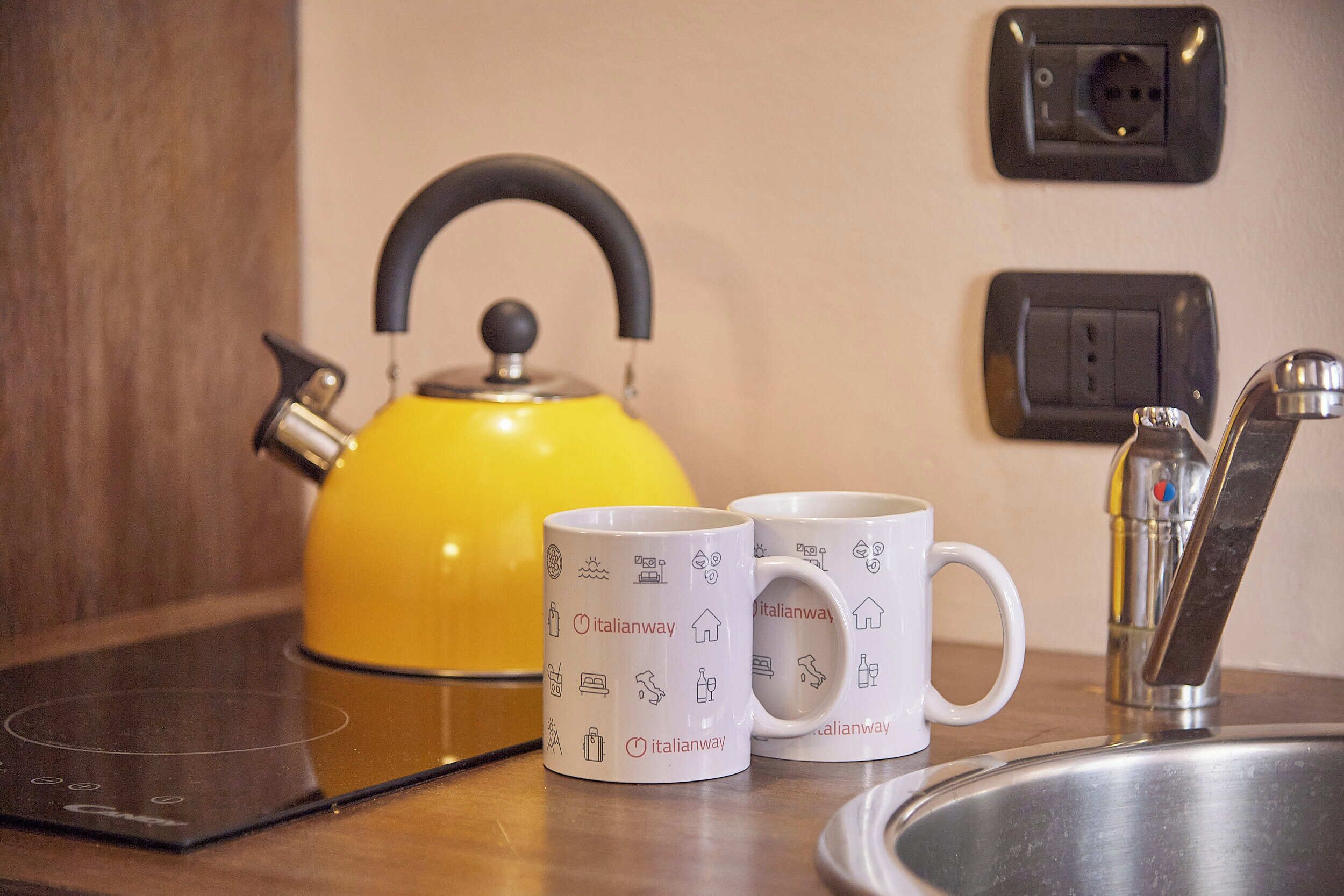Uma bancada de cozinha com uma chaleira amarela, duas canecas brancas com ícones relacionados a viagens e uma torneira moderna.