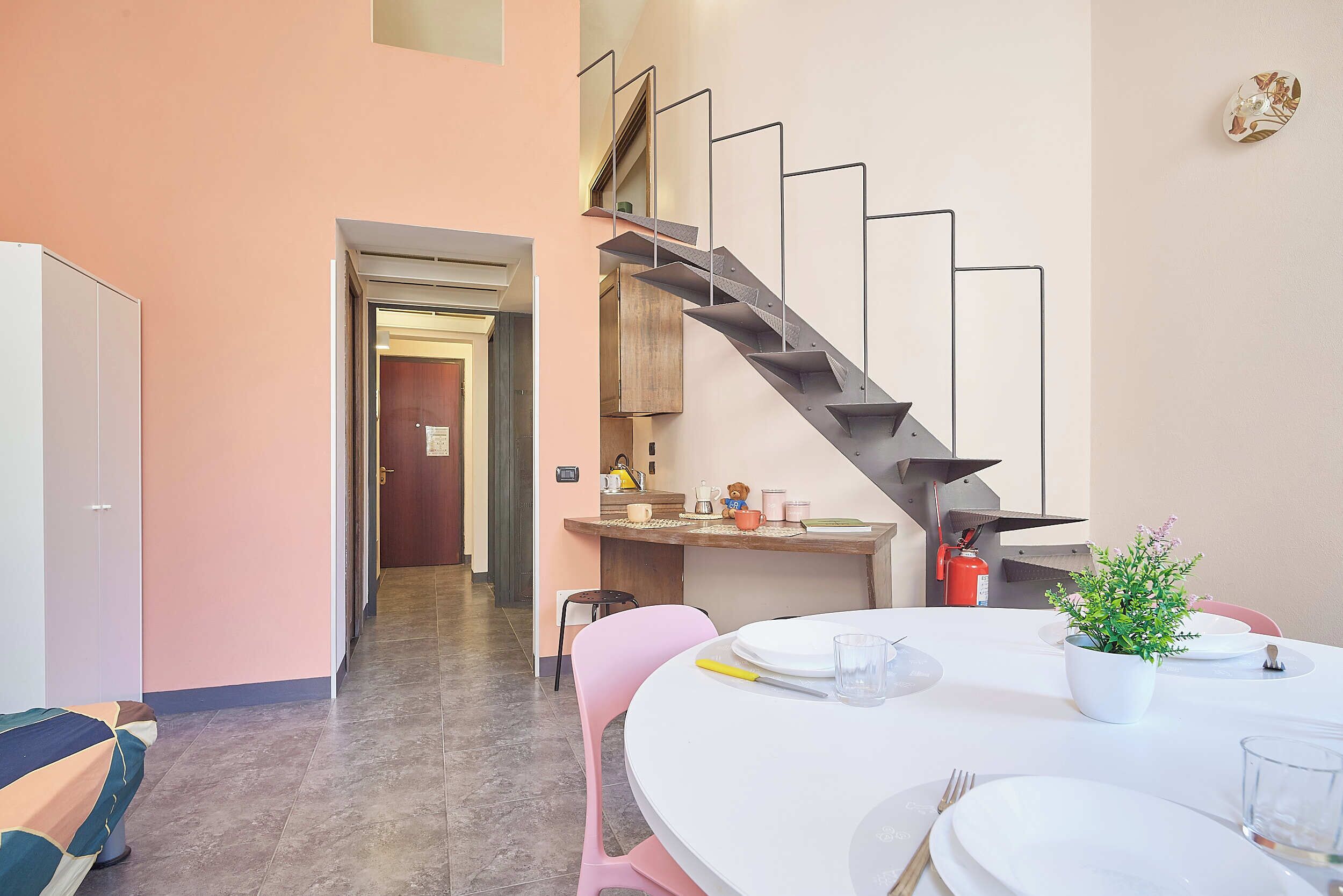A modern interior with a staircase, a dining area with a table set for two, and a kitchen area with a coffee machine.