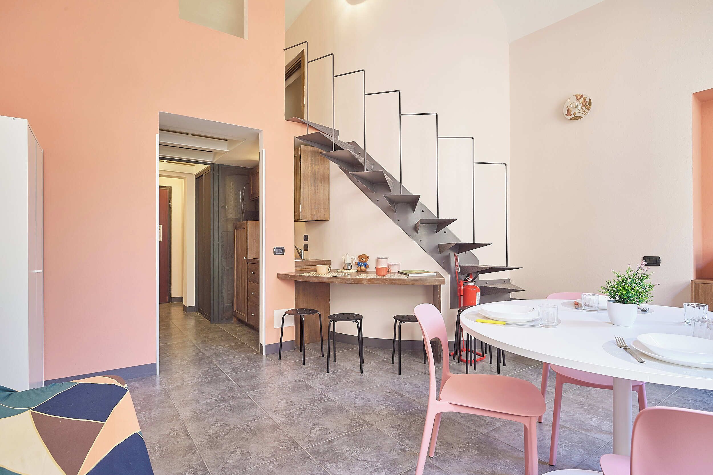 Una cocina moderna con estufa integrada, un comedor con sillas rosadas y una escalera que sube a otro nivel.