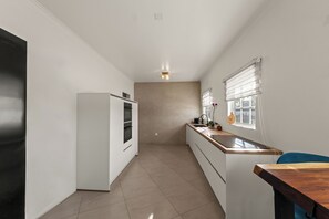 A narrow kitchen with white cabinets, a built-in oven, and a window with blinds. - Luxury Apartment Nestled in Beautiful Natural Surroundings (Willemstad)