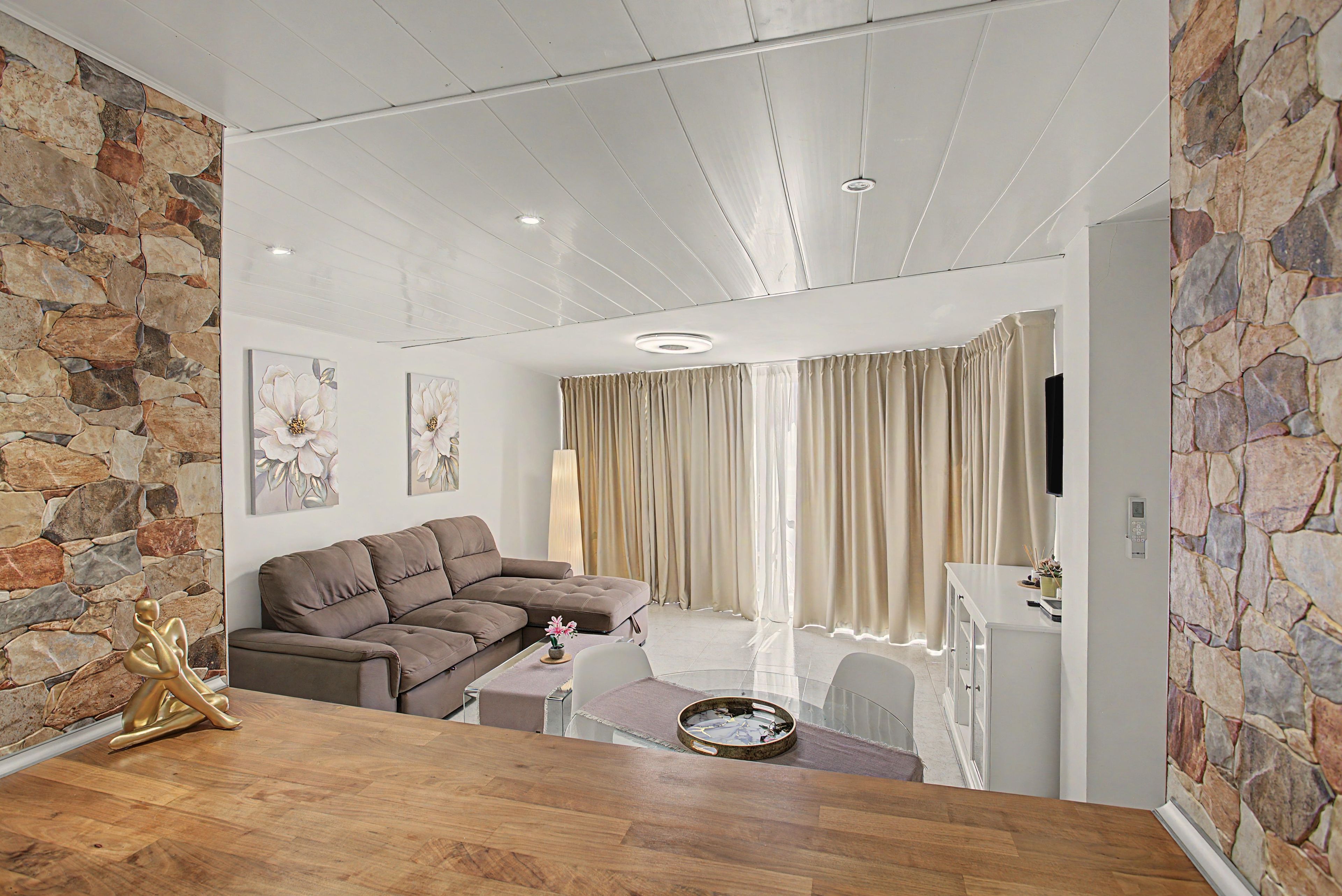 Een moderne woonkamer met een hoekbank, een eettafel met een bloemenvaas en een accentmuur van steen.