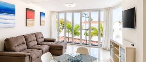A living room with a brown sofa, a dining table with a white chair, a TV mounted on the wall, and large windows with a view of greenery and buildings.