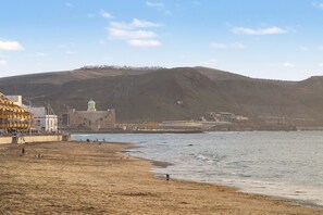 Uma área costeira com praia de areia, um edifício com estrutura em cúpula e paisagem montanhosa ao fundo.