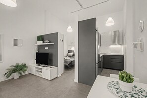 Ein moderner, minimalistischer Wohnraum mit Flachbildfernseher, einer kleinen Pflanze und einem Essbereich mit Tisch und Stühlen.