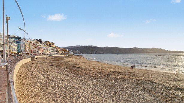 Una playa con un paseo marítimo, edificios y un paisaje montañoso al fondo.