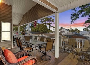 Eine überdachte Terrasse im Freien mit Tisch und Stühlen, Blick auf die Nachbarschaft und ein Sonnenuntergang im Hintergrund.