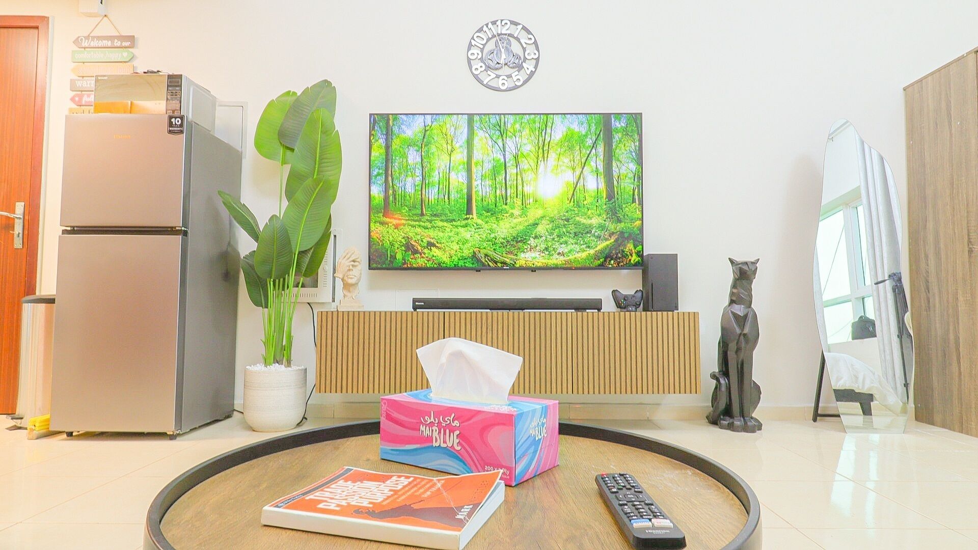 Un salon avec un téléviseur à écran plat, un réfrigérateur, une plante en pot, une télécommande et un livre posé sur une table ronde.
