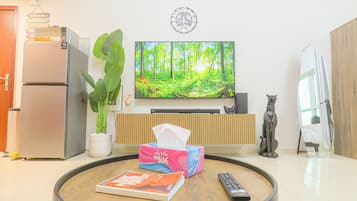 Un salon avec un téléviseur à écran plat, un réfrigérateur, une plante en pot, une télécommande et un livre posé sur une table ronde.