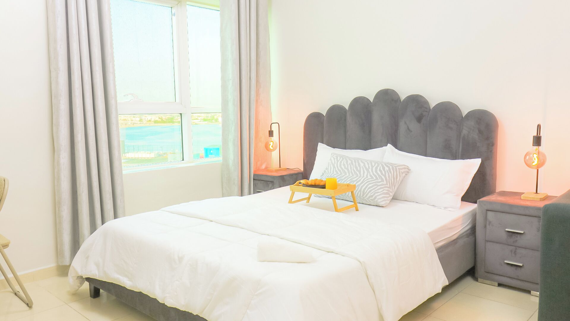 Een hotelkamer met een bed, een nachtkastje met een lamp, een stoel en een raam met gordijnen.