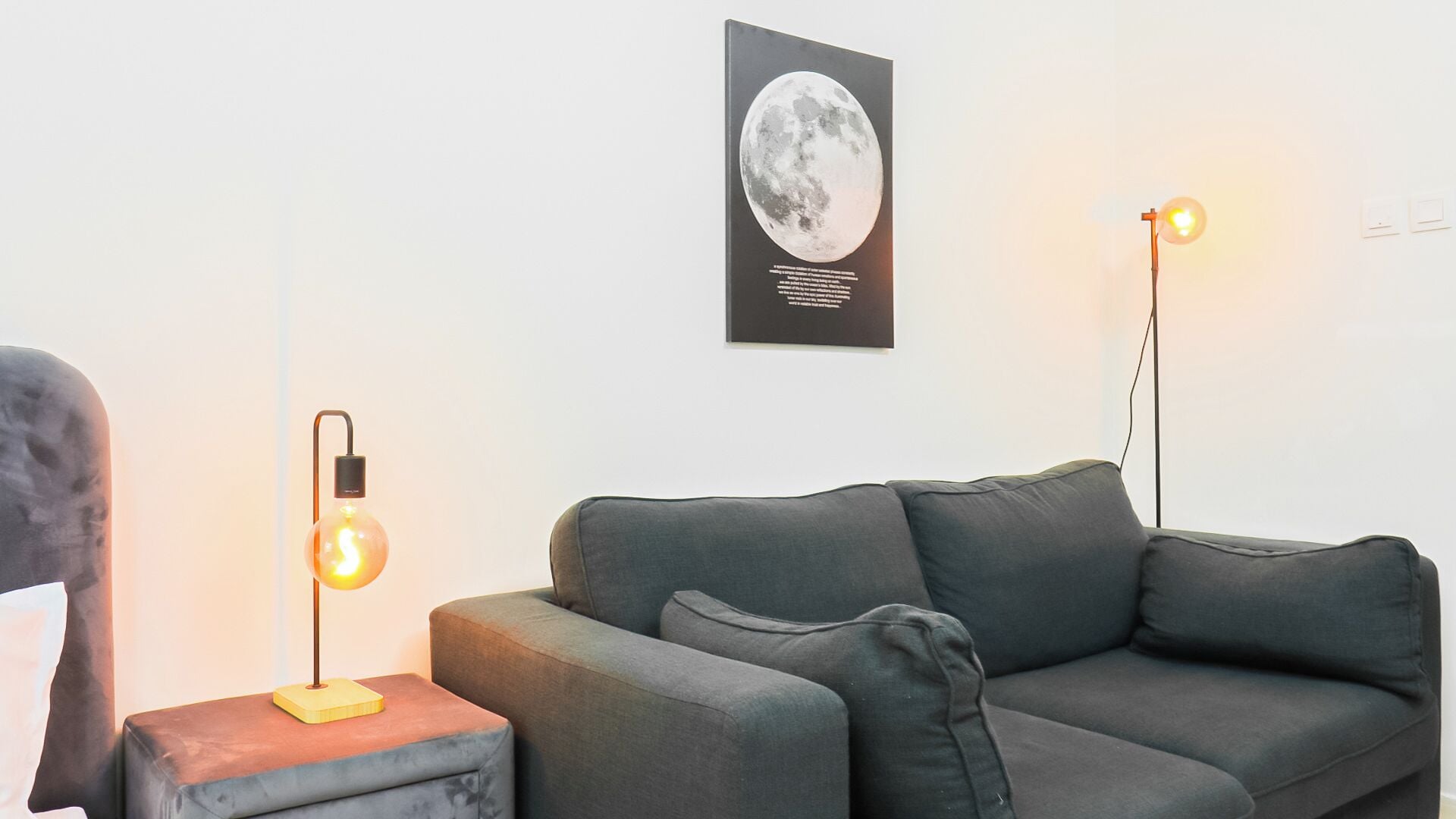 Un salon moderne avec un canapé gris, une petite table d’appoint et deux lampadaires. Un tableau encadré représentant la lune est accroché au mur.