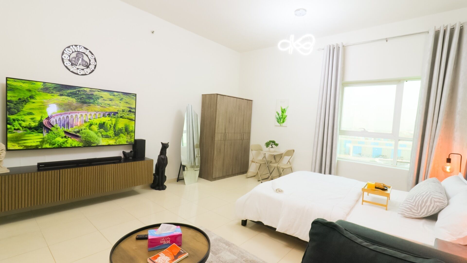 Ein modernes Hotelzimmer mit großem Flachbildfernseher, einem Bett, einem Sofa, einem kleinen Tisch, einem Schrank und einem Fenster mit Vorhängen.