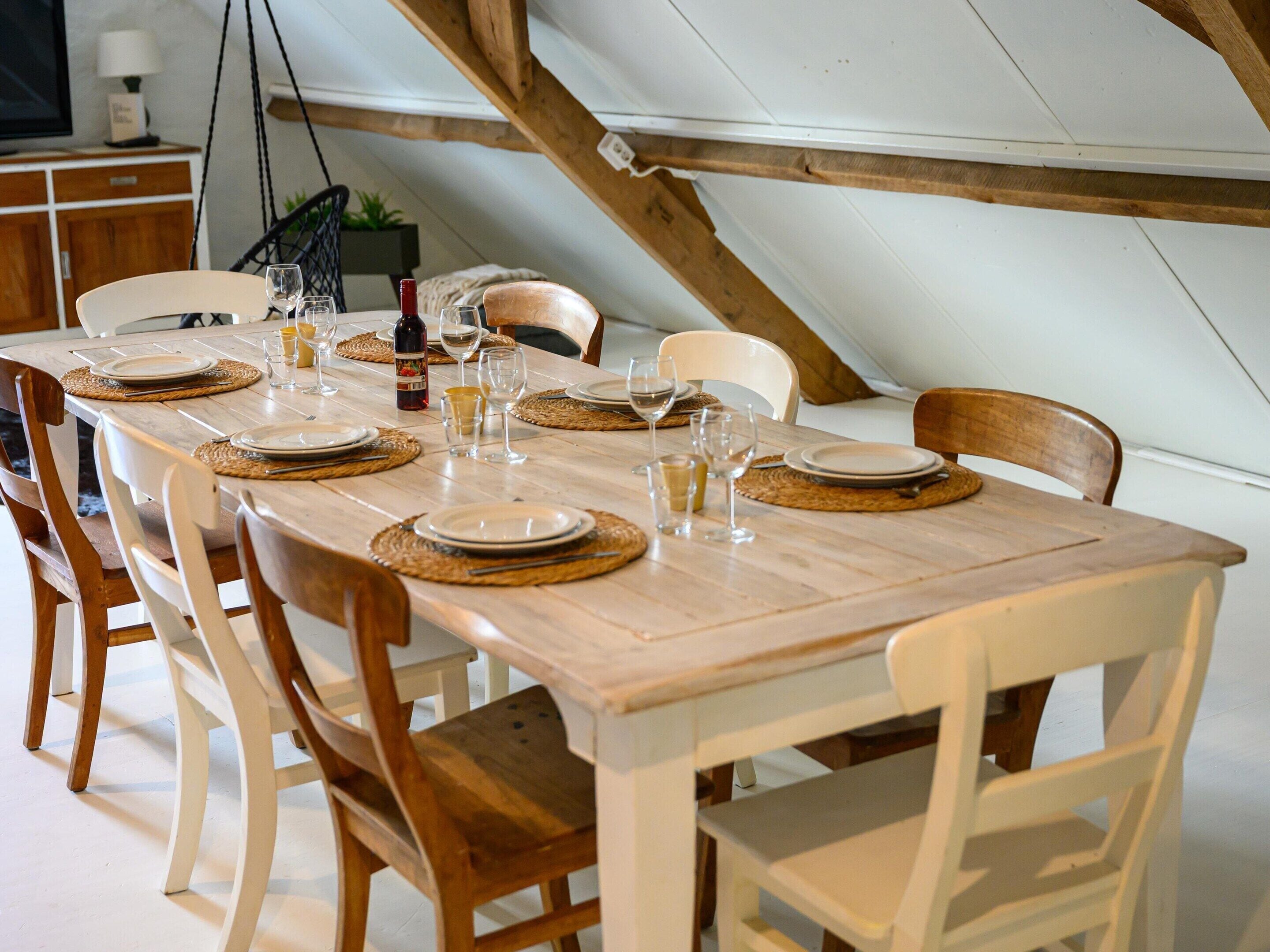 Une table à manger dressée avec des assiettes, des couverts et des verres, entourée de chaises en bois dans une pièce dont les poutres apparentes sont également en bois.