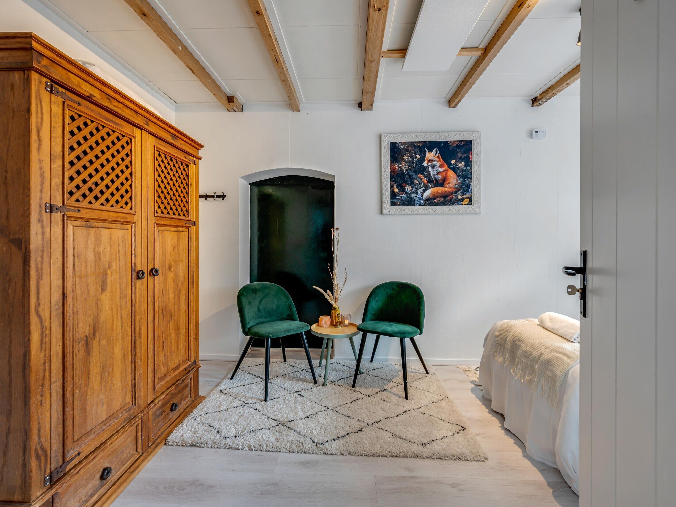 Une pièce avec une armoire en bois, deux chaises vertes, une petite table, un tableau au mur et un lit recouvert d’une couverture blanche.