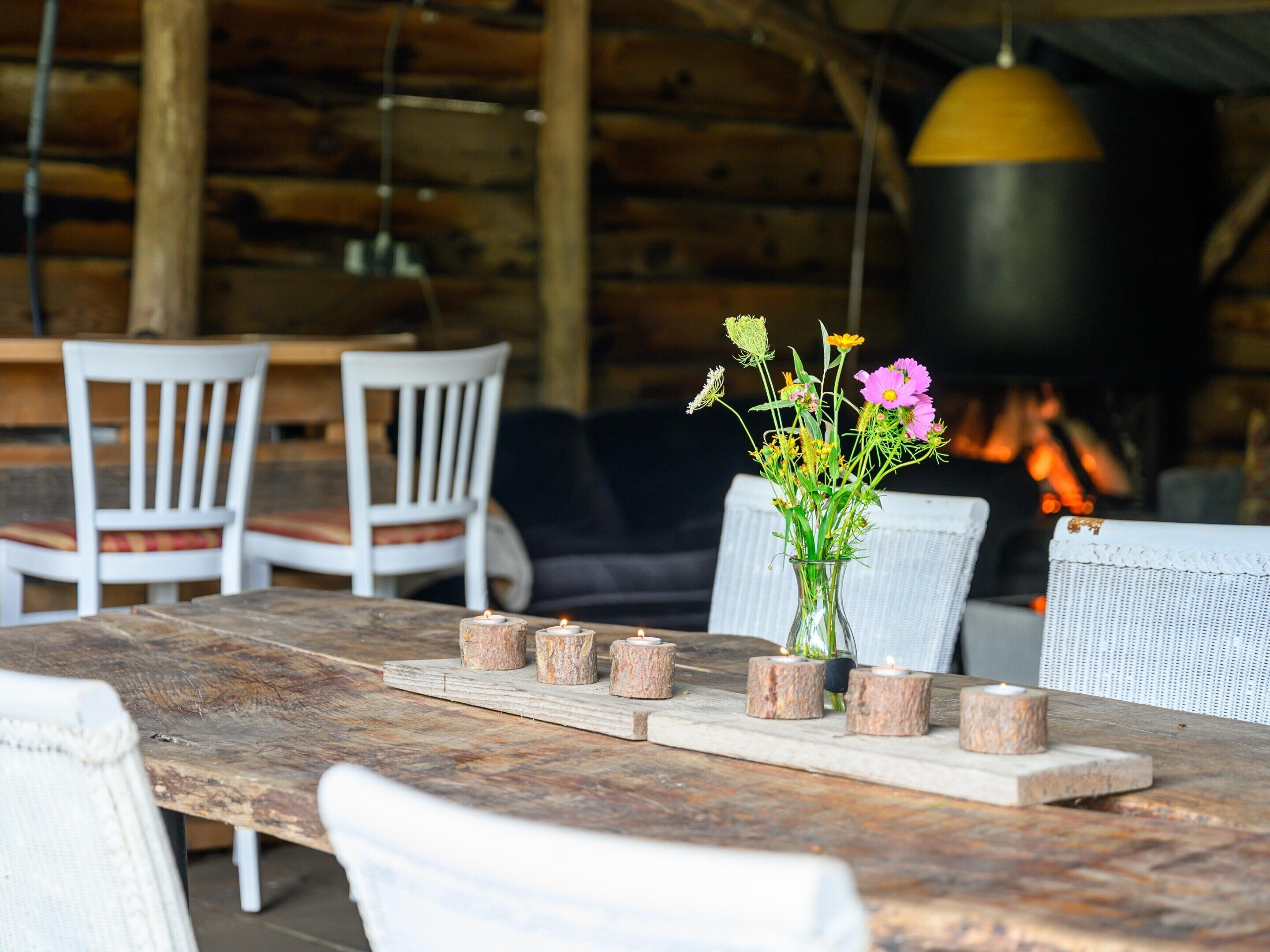 A rustic wooden table with candles and a vase of flowers, surrounded by white chairs in a cozy setting with a fireplace in the background.